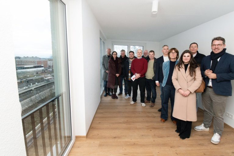 Eine Gruppe von Menschen steht in einer frisch gebauten und verputzten Wohnung und lächeln in die Kamera.