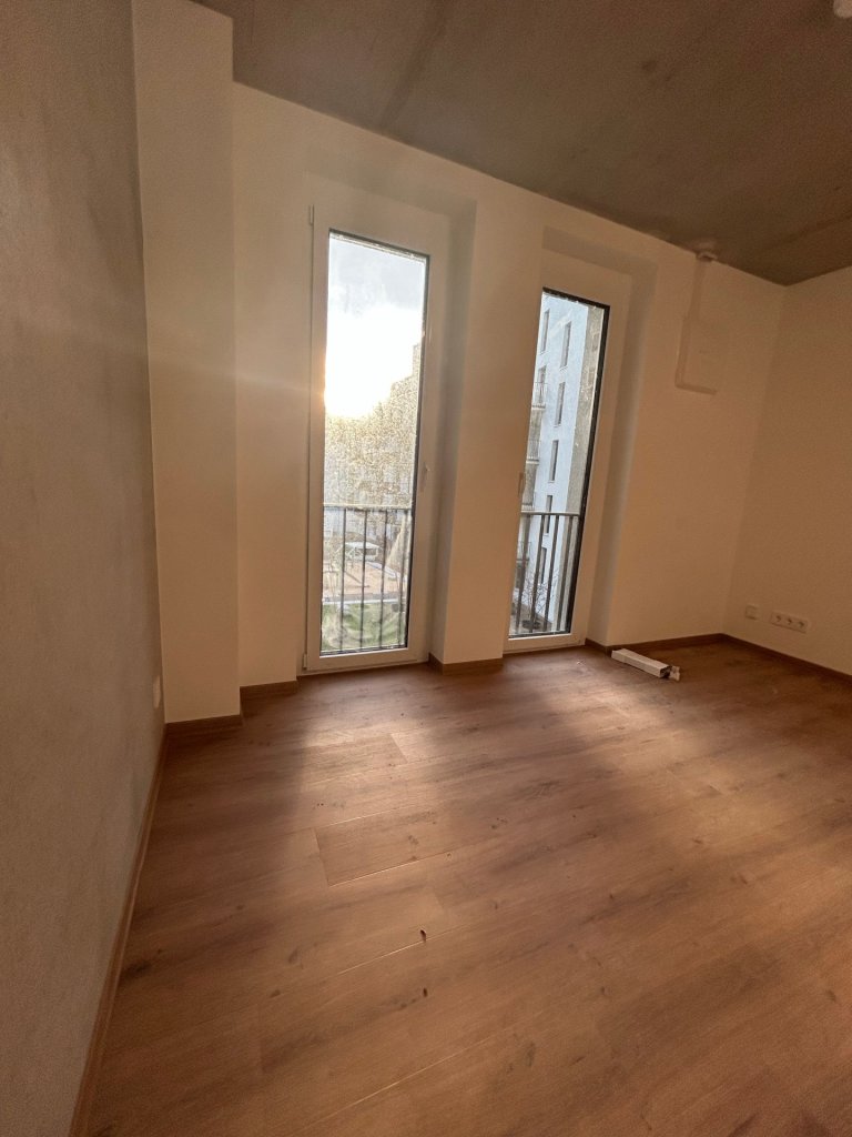 Eine frisch gebaute Wohnung. Durch zwei bodentiefe Fenster scheint Sonnenlicht