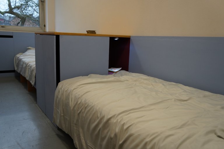 Zwei Betten stehen hintereinander in einem Raum mit Fenster. Sie werden miteinander von zwei hellblauen Schränken getrennt. In den Schränken ist eine kleine Nische als Ablagefläche integriert. 