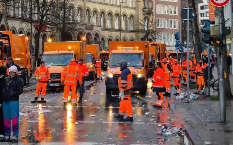 Viele Menschen mit Besen und Schaufeln in orangener Dienstkleidung laufen vor orangenen Einsatzfahrzeugen über eine Straße und reinigen diese von Müll.