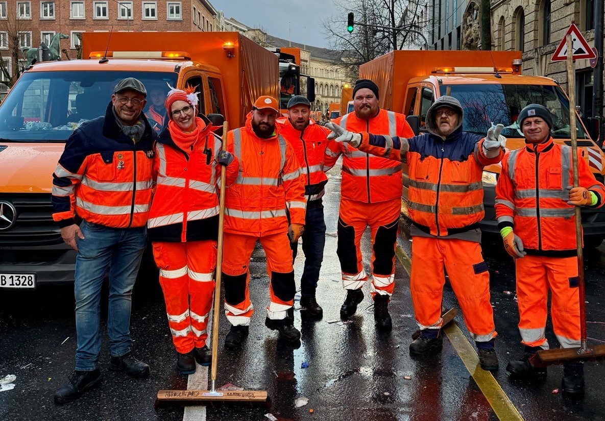 Sechs Menschen in orangener Dienstkleidung stehen Arm in Arm vor zwei orangenen Einsatzfahrzeugen und lächeln in die Kamera.