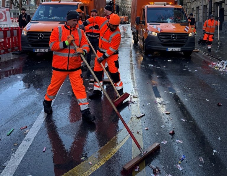 Zwei Männer mit Besen und Schaufeln in orangener Dienstkleidung laufen vor orangenen Einsatzfahrzeugen über eine Straße und reinigen diese von Müll.