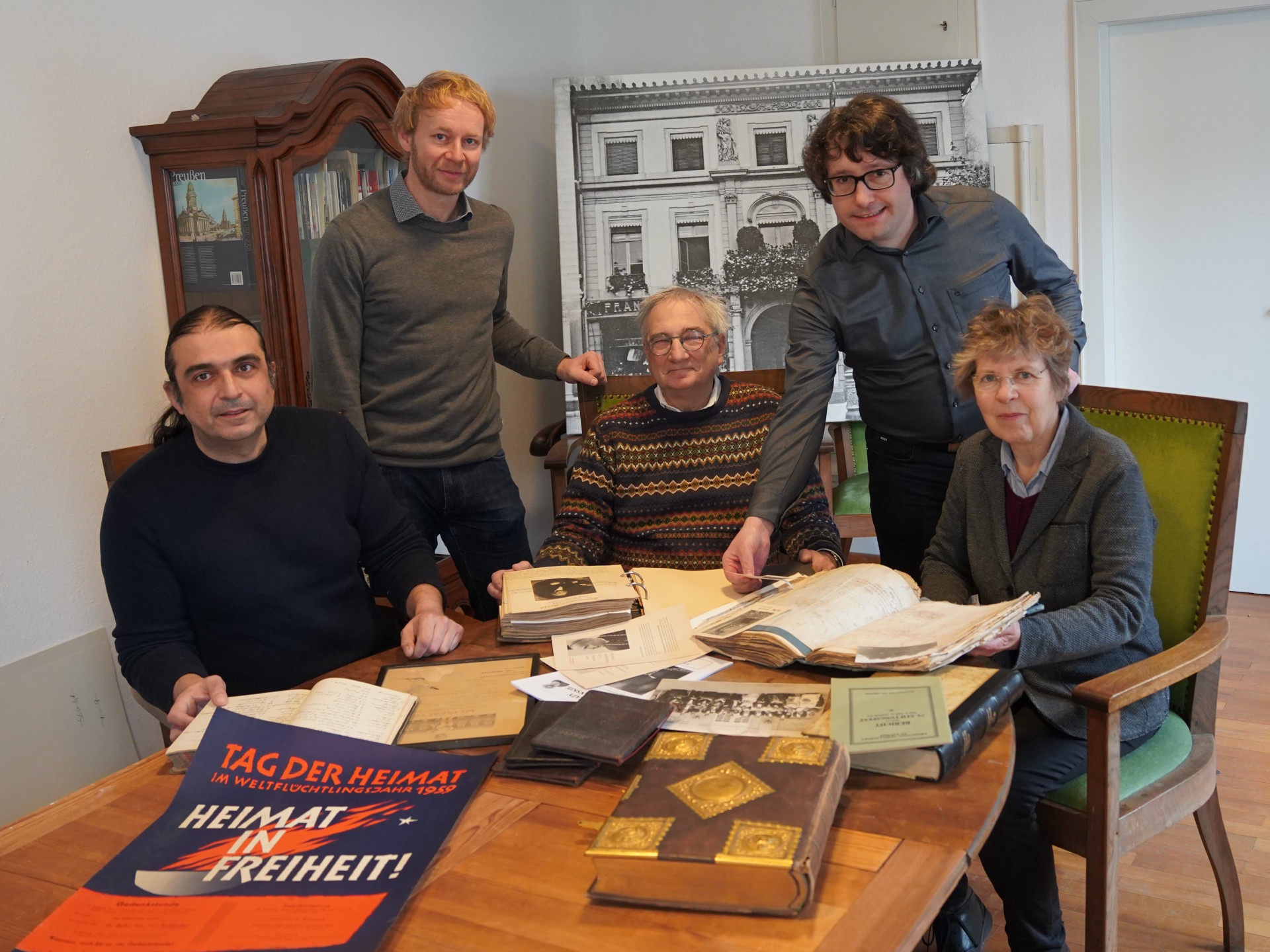 Fünf Menschen, drei sitzend, zwei stehend hinter einem Tisch, auf dem verschiedene Zeitdokumente, Listen, Plakate, Bücher liegen.