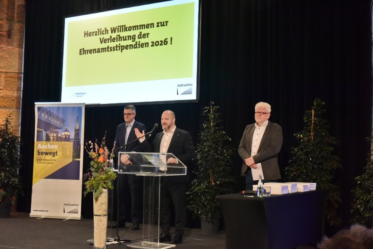 Ein Mann steht vor einem Podium mit Mikro, links und rechts von ihm stehen zwei weitere Männer.