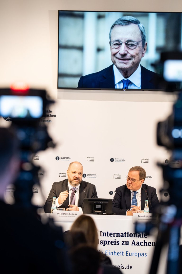 Zwei Männer sitzen auf einem Podium unter einem Bild eines anderen Mannes. 