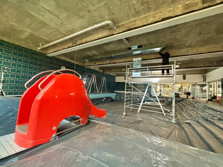 Ein Handwerker steht auf einem Gerüst in einem leeren Schwimmbecken. Im Vordergrund ist eine rote Kinderrutsche zu sehen.