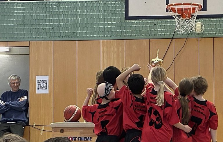 Kinder in roten Trikots heben unter einem Basketballkorb einen goldenen Pokal hoch.