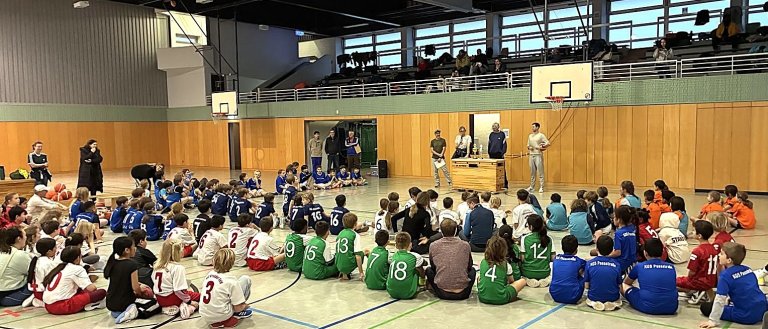 Kinder sitzen in einer Sporthalle und hören vier Erwachsenen zu, die vor zwei Pokalen stehen.