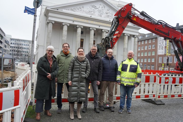 Eine Gruppe von sechs Menschen - zwei Frauen und vier Männer - steht vor einem Bagger am Theaterplatz