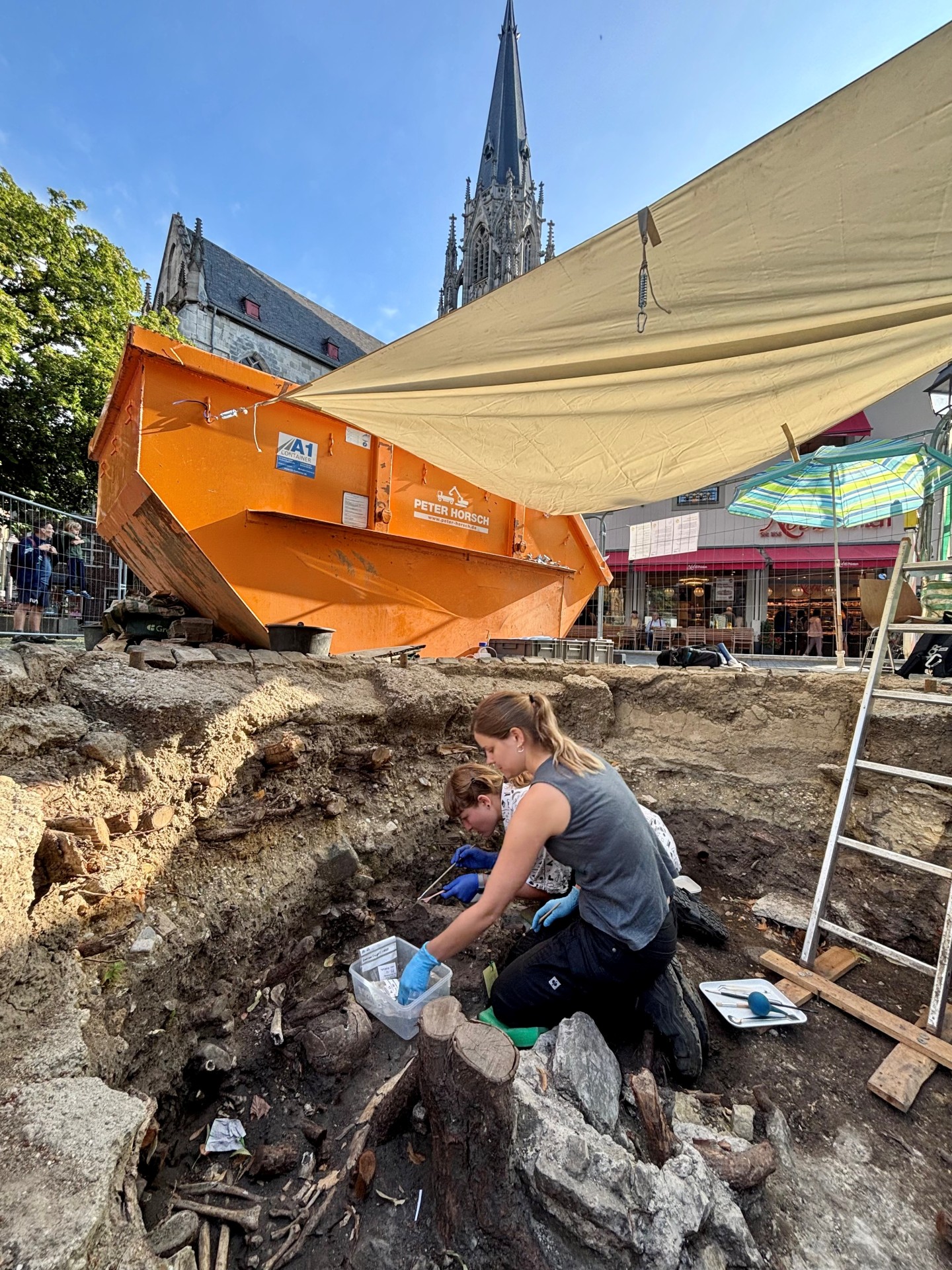 Zwei Archäologinnen in einer Grube, im Hintergrund ein Kirchturm.