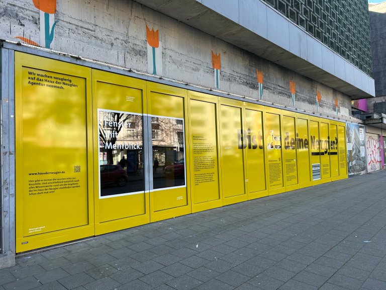 Eine beklebte Schaufensterfläche in Gelb mit schwarzer Schrift "Neugier?" und einem großen Sichtfenster.