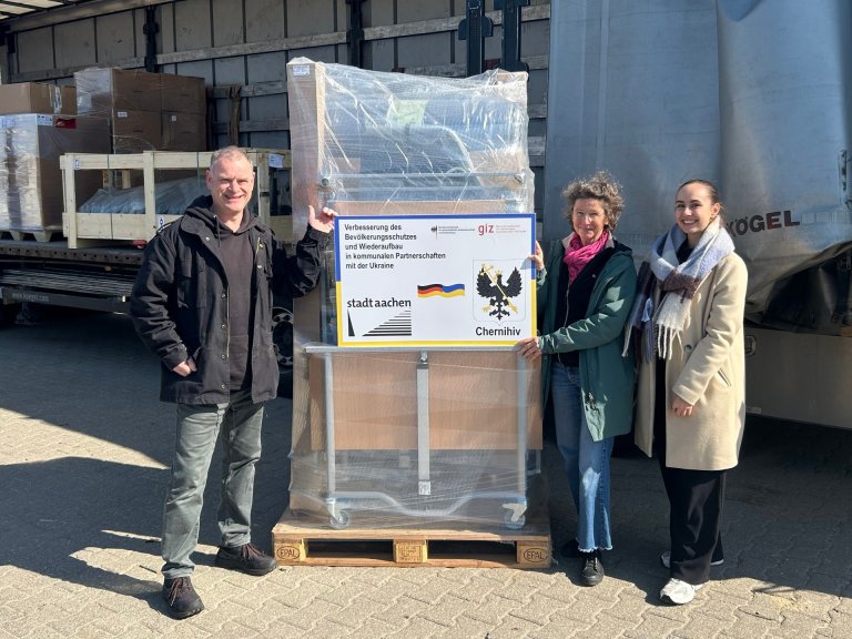 Drei Menschen stehen vor einem offenen LKW mit Hilfsgütern. Sie halten ein Schild mit den Logos der Städte Aachen und Chernihiv und dem Text: "Verbesserung des Bevölkerungsschutzes und Wiederaufbau in kommunalen Partnerschaften mit der Ukraine."