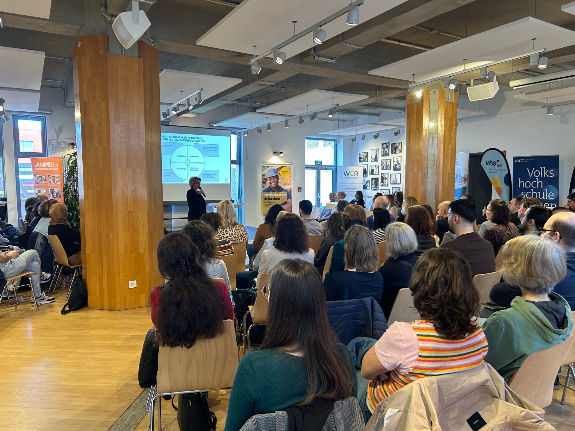 Viele Menschen sitzen um eine Bühne herum, auf der eine Frau einen Vortrag hält.