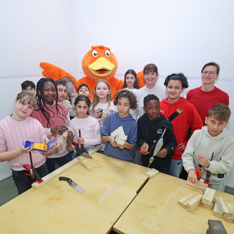 Eine Gruppe Kinder steht an einer Werkbank. Alle schauen in die Kamera, dahinter steht das Maskotchen der STAWAG, eine orangene Ente.