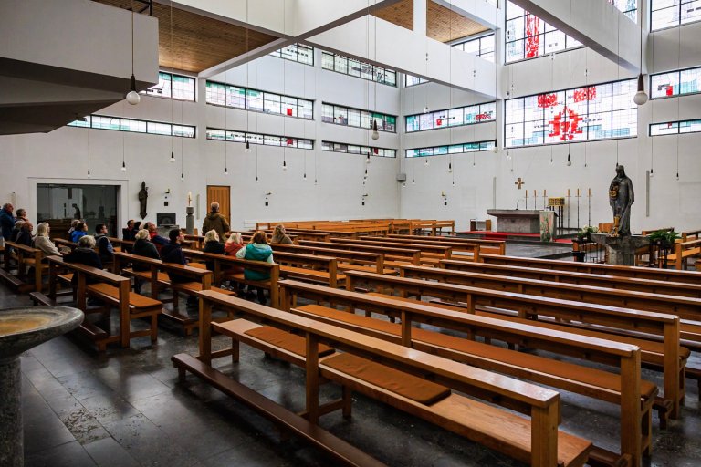 Ein moderner Kirchenraum von innen ist zu sehen; im Vordergrund stehen Holzbänke, im Hintergrund befinden sich einige Personen.