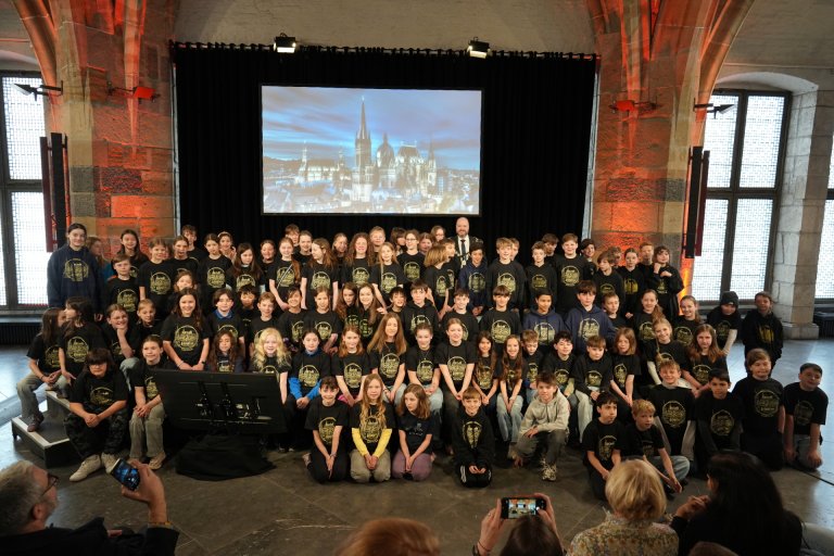 Eine große Gruppe Kinder in schwarzen T-Shirts sitzt und steht auf der Bühne im Aachener Krönungssaal. In der letzten Reihe steht Oberbürgermeister Michael Ziemons.