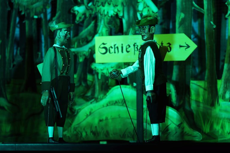 Zwei Stockpuppen - der Freischütz mit Gewehr um die Schultern und das Schängche neben ihm - stehen vor einem Schild "Schießplatz". Dahinter eine gemalte Kulisse mit Tannenwald.