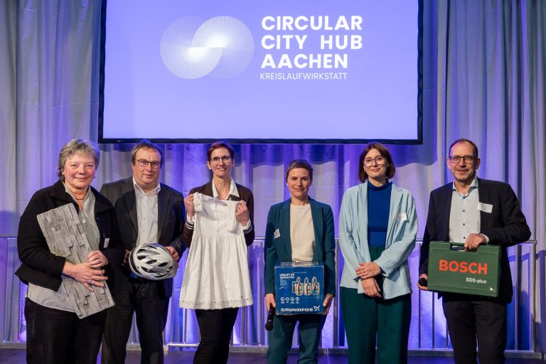 Sechs Personen - vier Frauen, zwei Männer - stehen nebeneinander auf einer Bühne. im Hintergrund steht auf einer großen Leinwand "Circular City Hub Aachen - Kreislaufwirtschaft".