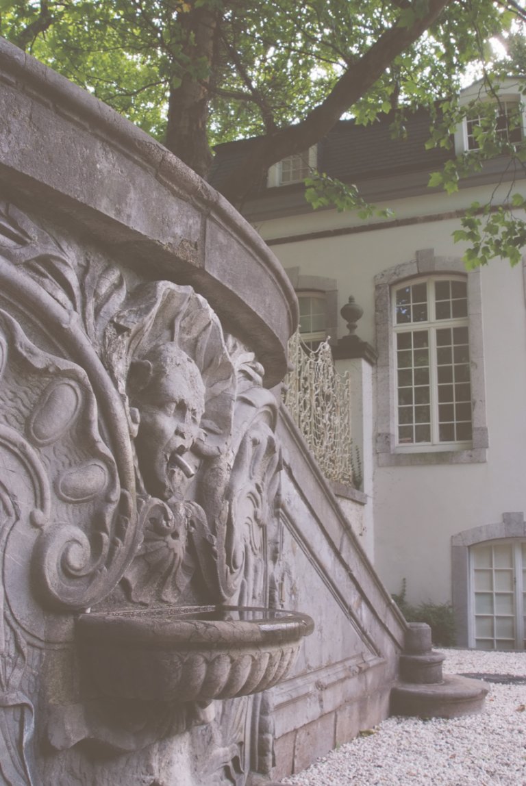 Im Vordergrund ist eine verzierte Stützmauer zu sehen. Im Hintergrund steht der Pavillon auf dem Lousberg.