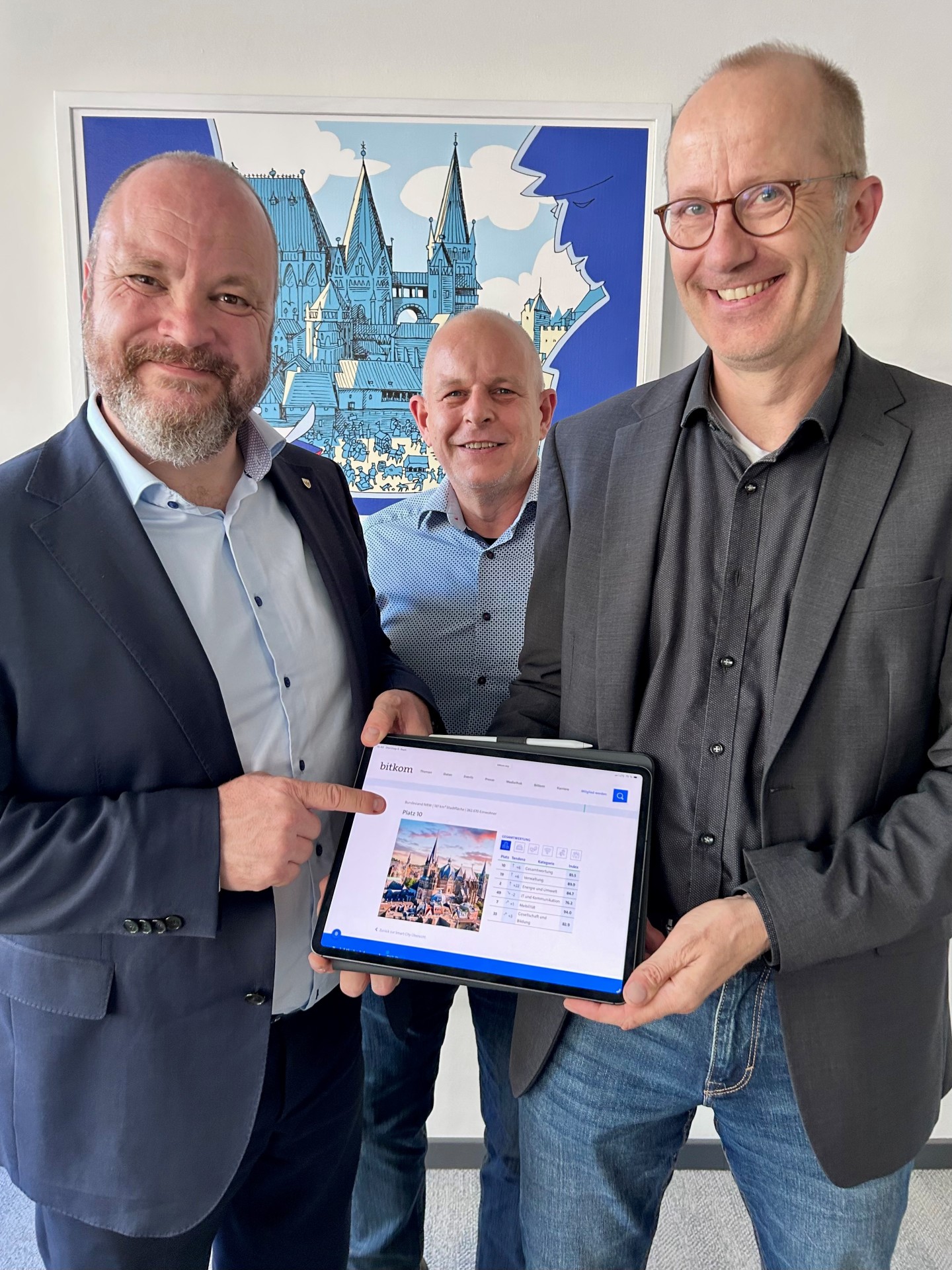 Drei Männer, darunter Aachens Oberbürgermeister, stehen beisammen und halten ein Tablet in der Hand.