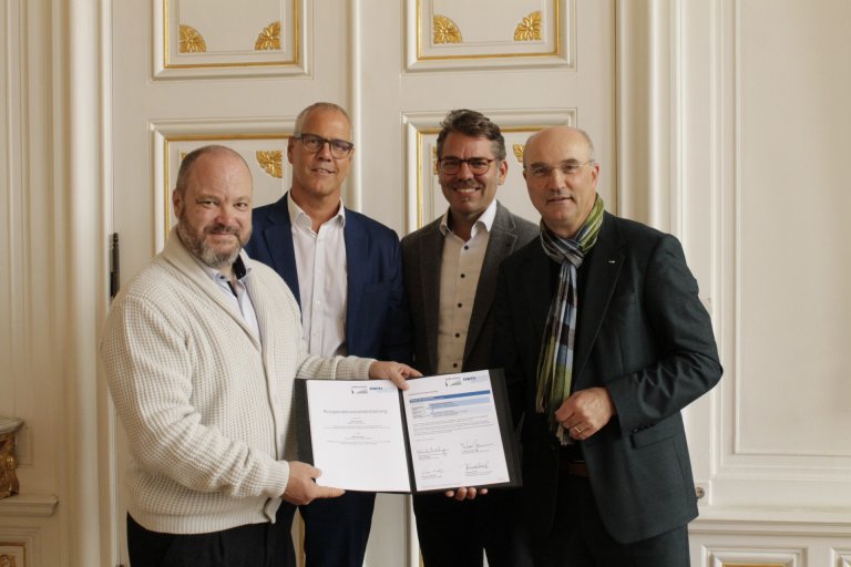 Vier Männer halten ein unterzeichnetes Dokument in den Händen und lächeln in die Kamera.