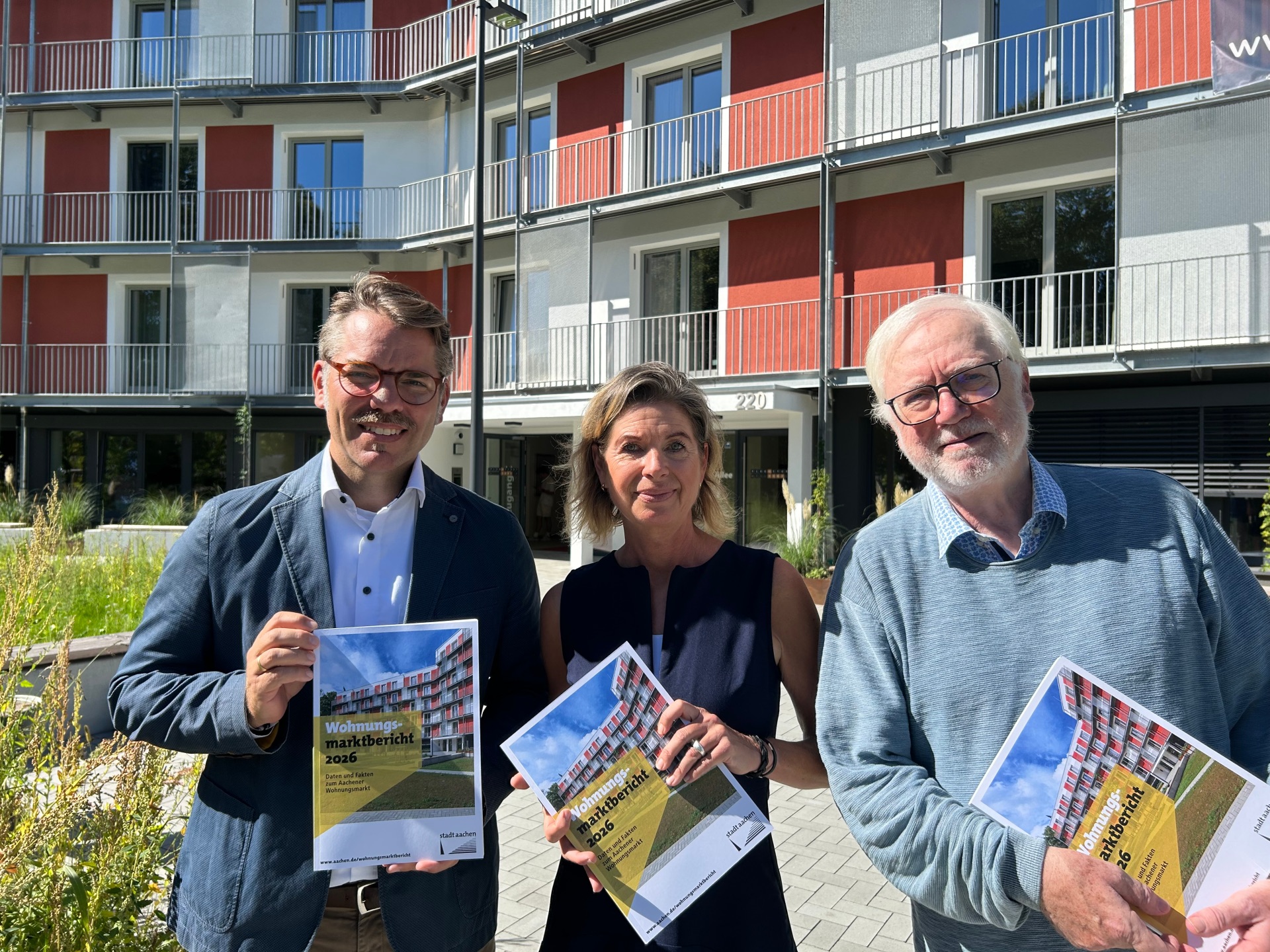 Zwei Männer und eine Frau stehen vor einem Neubaugebäude und halten Broschüren mit dem Titel "Wohnungsmarktbericht 2026" in den Händen.