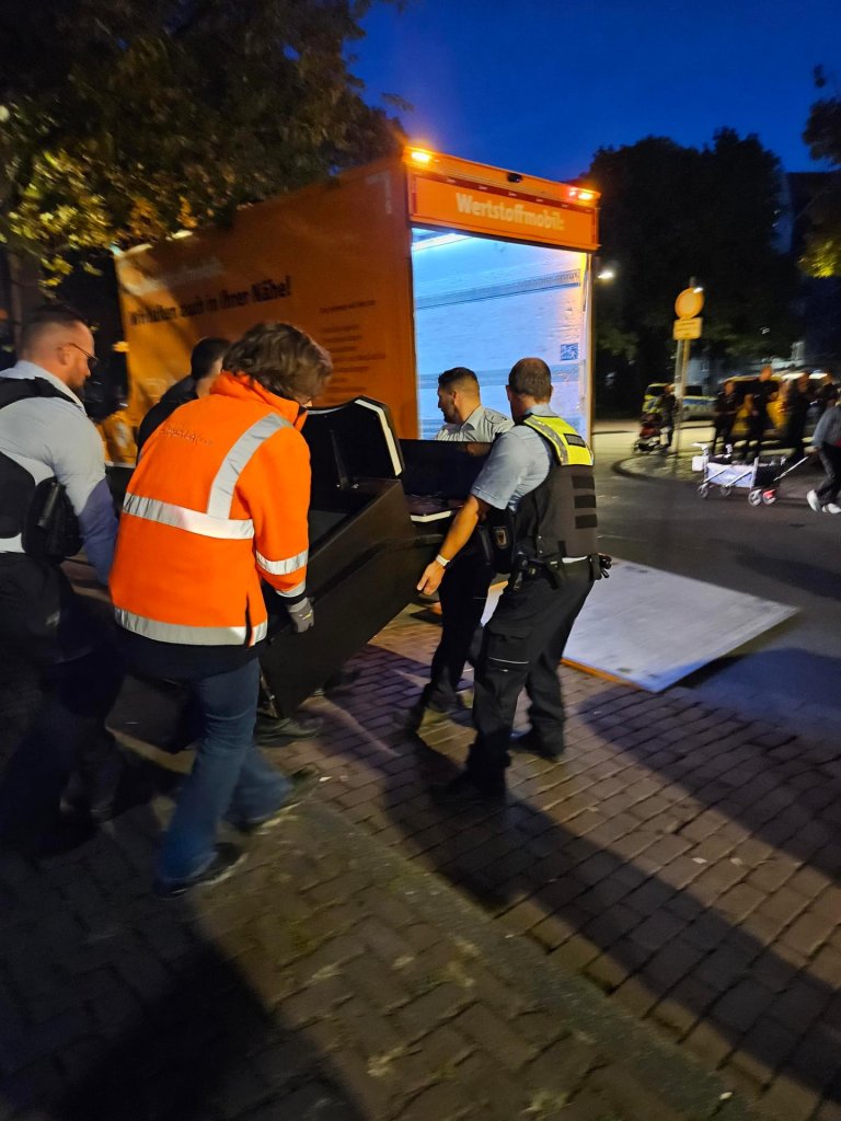 Vier Menschen tragen einen Spielautomaten zu einem Transporter.
